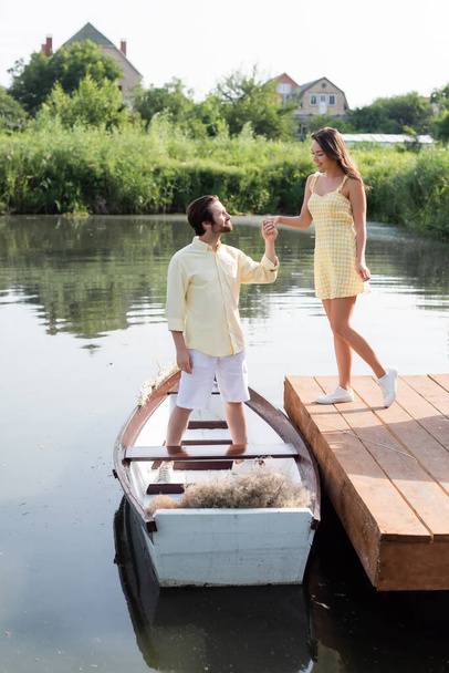vue latérale de l'homme barbu heureux en vêtements d'été tenant la main avec petite amie gaie près du bateau - Photo, image