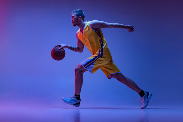 Studio shot of muscled man, basketball player training with ball isolated on purple background in neon light. Goals, sport, motion, activity concepts. - Фото, изображение