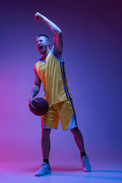 Studio shot of muscled man, basketball player training with ball isolated on purple background in neon light. Goals, sport, motion, activity concepts. - Photo, Image