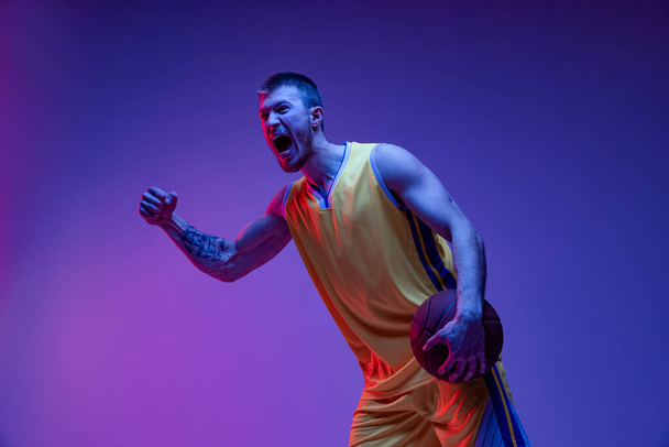Studio shot of muscled man, basketball player training with ball isolated on purple background in neon light. Goals, sport, motion, activity concepts. - Foto, Imagen