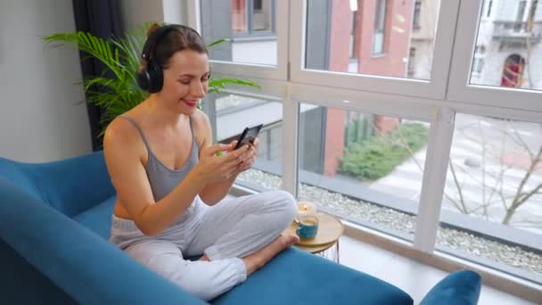 Vrouw in draadloze hoofdtelefoon die naar muziek luistert en mobiele apps gebruikt of communiceert op sociale netwerken op een smartphone die thuis op de bank zit - Video