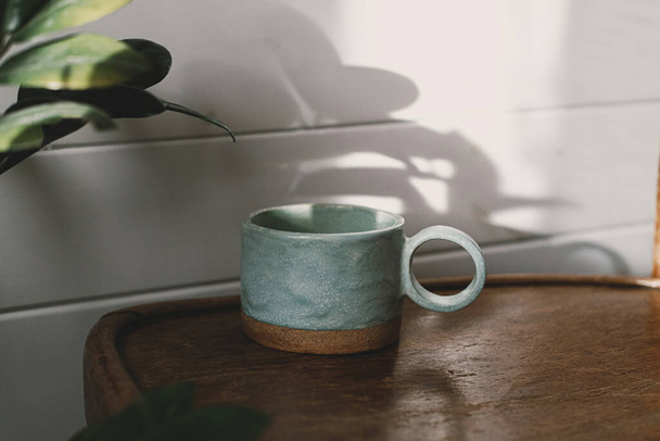 Stilvolle Tasse in Sonnenlicht und Schatten auf rustikalem Hintergrund. Moderne Keramik minimalistische Tasse auf Holzstuhl in stilvollen sonnigen Boho-Raum mit Pflanzen. Ästhetik des Morgenkaffees - Foto, Bild