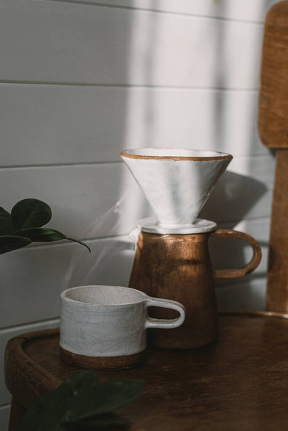 Moderne Keramiktasse, Wasserkocher und Tropfer zum Aufgießen von Kaffee im Sonnenlicht auf rustikalem Holzstuhl und hinterlässt Schatten in stilvollem, sonnigem Raum. Alternative Kaffeebrühmethode. - Foto, Bild