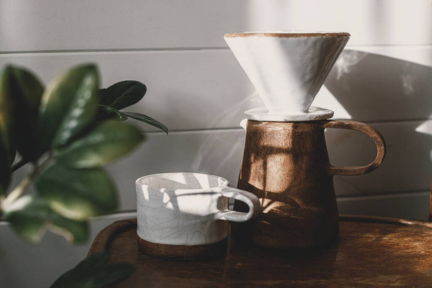 Ranní kávová estetika. Moderní keramický šálek, konvice a kapátko na přelévání kávy ve slunečním světle na venkovské dřevěné židli a zanechává stín ve stylové slunné místnosti. Alternativní metoda vaření kávy - Fotografie, Obrázek