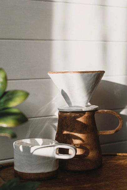 Alternative Kaffeebrühmethode. Moderne Keramiktasse, Wasserkocher und Tropfer zum Aufgießen von Kaffee im Sonnenlicht auf rustikalem Holzstuhl und hinterlässt Schatten in stilvollem, sonnigem Raum - Foto, Bild