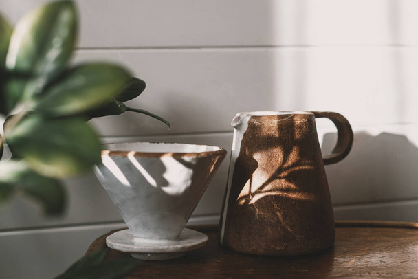 Morgenkaffee-Ästhetik. Moderne Keramiktasse, Wasserkocher und Tropfer zum Aufgießen von Kaffee im Sonnenlicht auf rustikalem Holzstuhl und hinterlässt Schatten in stilvollem, sonnigem Raum. Alternative Kaffeebrühmethode - Foto, Bild
