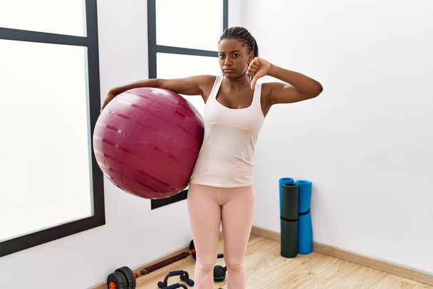 Young african american woman with braids holding pilate ball at yoga room with angry face, negative sign showing dislike with thumbs down, rejection concept  - Foto, Imagem