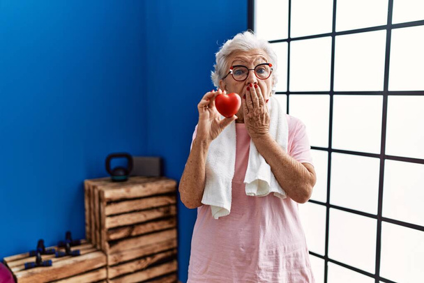 Mujer mayor con pelo gris usando ropa deportiva sosteniendo el corazón rojo cubriendo la boca con la mano, conmocionada y temerosa de equivocarse. expresión sorprendida  - Foto, imagen