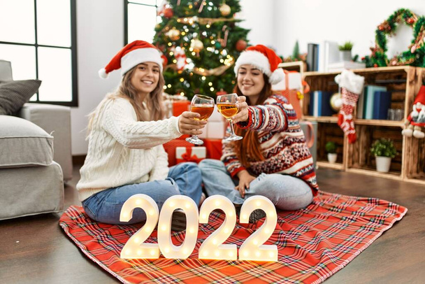 Woman couple toasting with champagne for new year 2022 sitting by christmas tree at home - Photo, Image