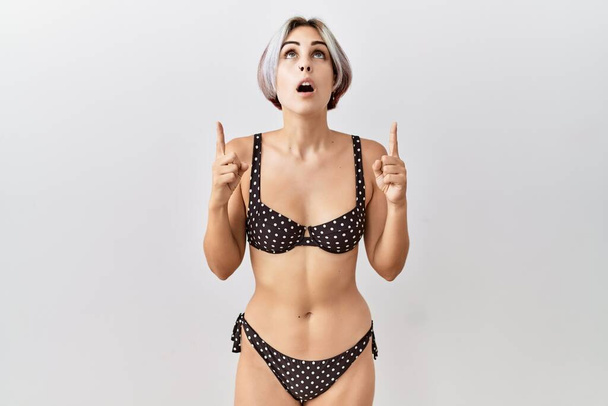 Young beautiful woman wearing swimsuit over isolated background amazed and surprised looking up and pointing with fingers and raised arms.  - Photo, Image