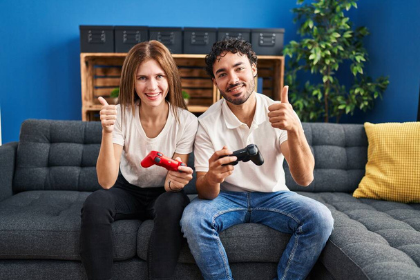 Joven pareja jugando videojuego celebración controlador en casa sonriendo feliz y positivo, pulgar hacia arriba haciendo excelente y signo de aprobación  - Foto, Imagen