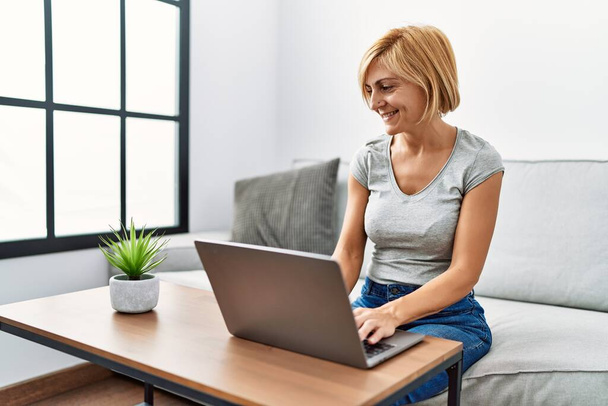 Блондинка середнього віку посміхається впевнено використовуючи ноутбук вдома
 - Фото, зображення
