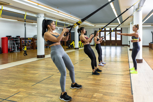 Entrenadora que explica el ejercicio a las deportistas que cuelgan de las cuerdas TRX durante el entrenamiento de suspensión en un amplio gimnasio ligero - Foto, Imagen