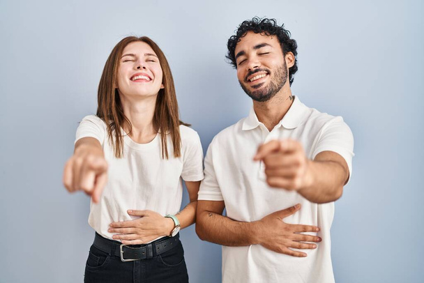 Jeune couple portant des vêtements décontractés debout ensemble riant de vous, pointant du doigt la caméra avec la main sur le corps, expression de honte  - Photo, image