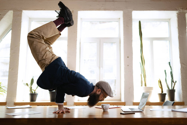 Jeune homme barbu, employé de bureau s'amusant, faisant du yoga sur une table en bois dans un bureau moderne au travail avec des gadgets. Concept d'entreprise, mode de vie sain, sport, passe-temps - Photo, image