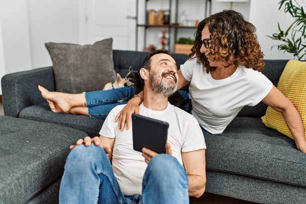Ein hispanisches Paar mittleren Alters lächelt glücklich und benutzt ein Touchpad. Mit Hunden zu Hause auf dem Sofa sitzen. - Foto, Bild