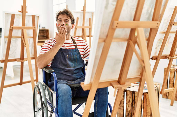 Homem bonito jovem com barba no estúdio de arte sentado em cadeira de rodas cobrindo a boca com a mão, chocado e com medo de erro. expressão surpreendida  - Foto, Imagem