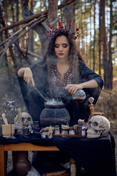 Witch in black, long dress, with red crown in her long hair. Posing in pine forest. Making a magic potion. Spells and witchcraft. Close-up, smoke. - Fotoğraf, Görsel