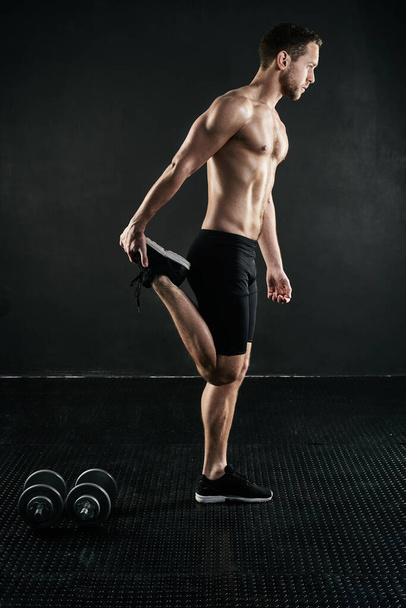 De pre-workout stretch. Studio shot van een shirtloze jonge man opwarmen tegen een donkere achtergrond. - Foto, afbeelding