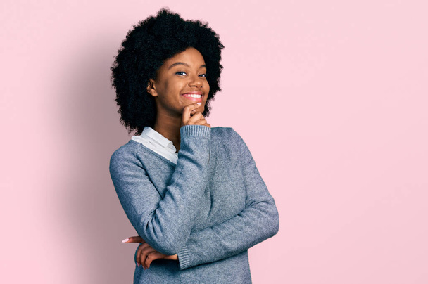 Νεαρή Αφροαμερικανή γυναίκα που φοράει επαγγελματικά ρούχα και κοιτάζει με αυτοπεποίθηση την κάμερα με χαμόγελο με σταυρωμένα τα χέρια και το χέρι υψωμένο στο πηγούνι. θετική σκέψη.  - Φωτογραφία, εικόνα