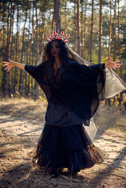 Heks in zwarte, lange jurk, met rode kroon in haar lange haar onder een zwarte sluier. Poseren in het dennenbos. Spreuken, magie en hekserij. Totale lengte. - Foto, afbeelding