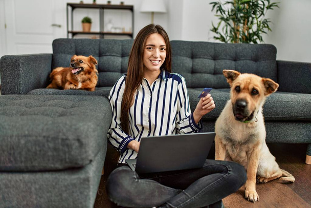 Young hispanic woman using laptop and credit card sitting on floor with dogs at home - Photo, image