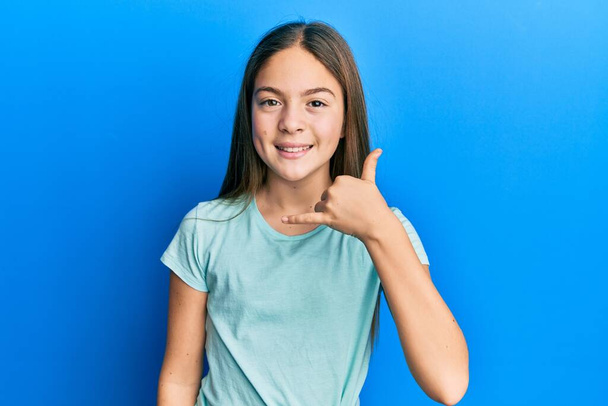 Красивая брюнетка маленькая девочка в обычной белой футболке улыбается делая телефонный жест с рукой и пальцами, как говорить по телефону. коммуникационные концепции.  - Фото, изображение