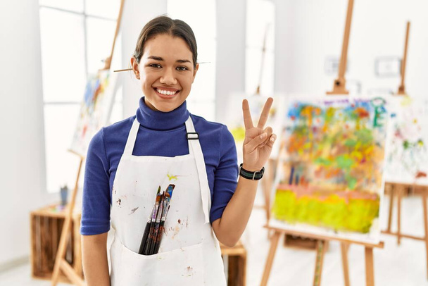 Nuori brunette nainen art studio näyttää ja osoittaa sormilla numero kaksi hymyillen itsevarma ja onnellinen.  - Valokuva, kuva