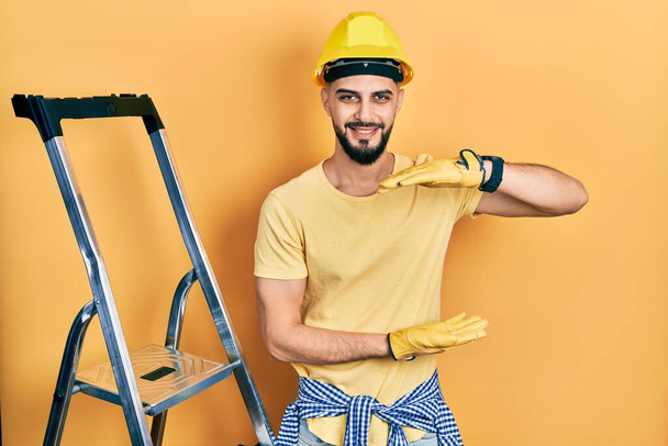 Handsome man with beard by construction stairs wearing hardhat gesturing with hands showing big and large size sign, measure symbol. smiling looking at the camera. measuring concept.  - Photo, Image