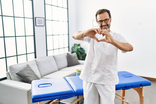 Középkorú fizioterápiás férfi dolgozik otthon mosolyogva a szerelemben mutató szív szimbólum és alakja kézzel. romantikus koncepció.  - Fotó, kép
