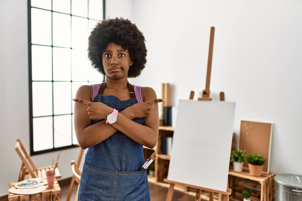 Jovem afro-americana com cabelo afro no estúdio de arte apontando para ambos os lados com os dedos, direção diferente discordo  - Foto, Imagem