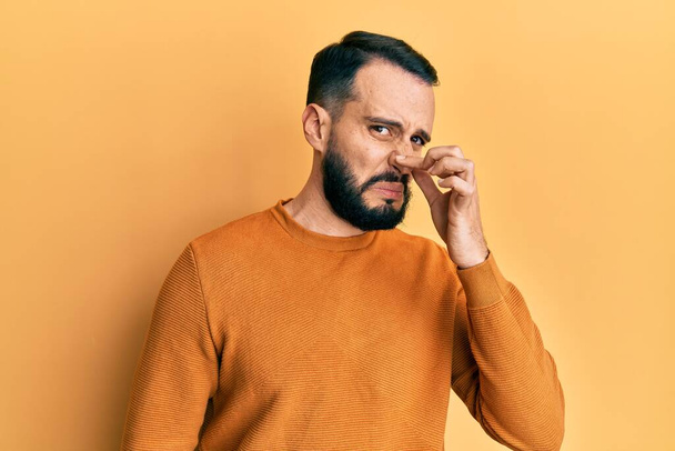 Young man with beard wearing casual winter sweater smelling something stinky and disgusting, intolerable smell, holding breath with fingers on nose. bad smell  - Foto, immagini