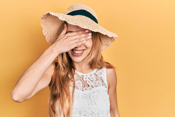 Mujer caucásica joven con sombrero de verano sonriendo y riendo con la mano en la cara cubriendo los ojos para sorpresa. concepto ciego.  - Foto, imagen