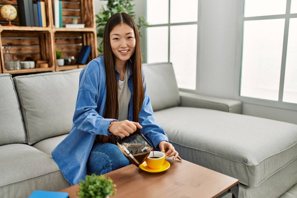 Genç Çinli kız evdeki kanepede oturup kahve içiyor.. - Fotoğraf, Görsel