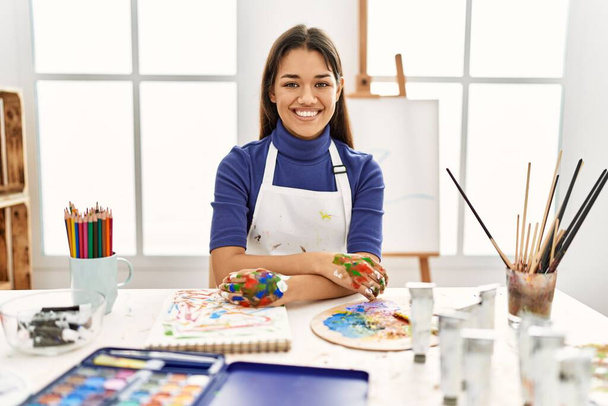 Νεαρή μελαχρινή γυναίκα σε στούντιο τέχνης με ζωγραφισμένα χέρια χαρούμενο πρόσωπο χαμογελώντας με σταυρωμένα χέρια κοιτάζοντας την κάμερα. θετικό πρόσωπο.  - Φωτογραφία, εικόνα