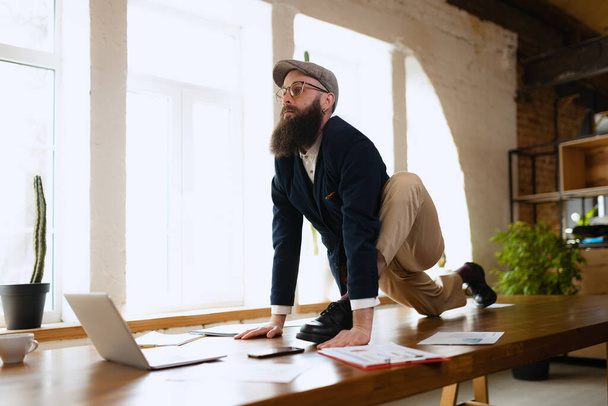 Young bearded man, office clerk having fun, doing yoga on wooden table in modern office at work time with gadgets. Concept of business, healthy lifestyle, sport, hobby - Photo, Image
