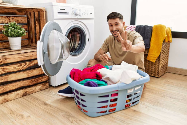 Young handsome man putting dirty laundry into washing machine smiling looking to the camera showing fingers doing victory sign. number two.  - Foto, immagini