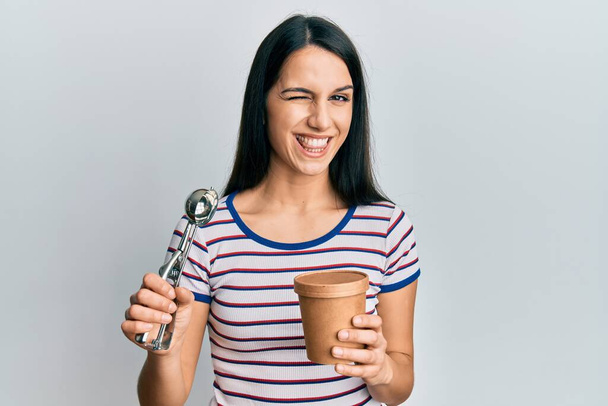 Joven mujer hispana sosteniendo helado guiñando el ojo mirando a la cámara con expresión sexy, cara alegre y feliz.  - Foto, Imagen