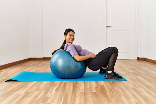 Mujer latina joven haciendo ejercicios abdominales usando la pelota en forma en el centro deportivo - Foto, Imagen