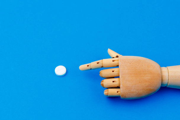 Ένα ξύλινο χέρι δείχνει ένα κλασικό ιατρικό λευκό χάπι με βαθμολογία σε ένα παραδοσιακό μπλε φόντο. Επιλεκτική εστίαση στο χάπι με χώρο αντίγραφο για το κείμενο - Φωτογραφία, εικόνα