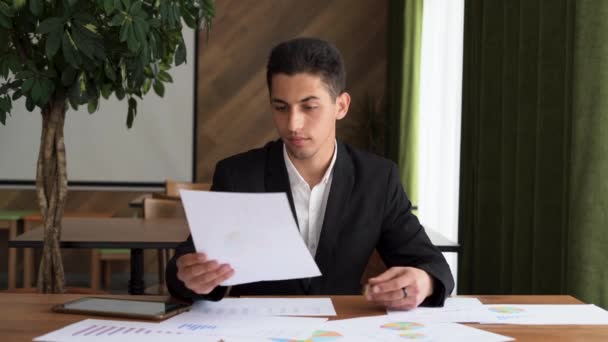 Hombre de negocios árabe joven concentrado mirando a través de informe de investigación de marketing, analizando datos estadísticos en gráficos, desarrollando estrategia de crecimiento, trabajando solo en la oficina moderna - Metraje, vídeo