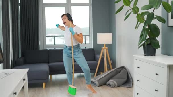 Γυναίκα με ακουστικά καθαρίζει το σπίτι και να διασκεδάσουν τραγουδώντας με μια σκούπα σαν αστέρι σε μια συναυλία - Πλάνα, βίντεο