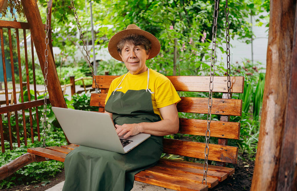 Senior woman gardener in a hat is watching something on a laptop computer and making a video call in the yard while outdoors. - Φωτογραφία, εικόνα