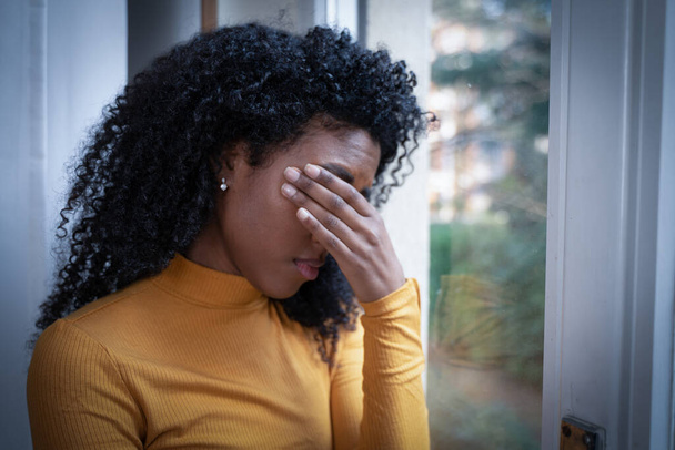 Μια μαύρη γυναίκα πολύ λυπημένη να σκέφτεται μπροστά στο παράθυρο. - Φωτογραφία, εικόνα