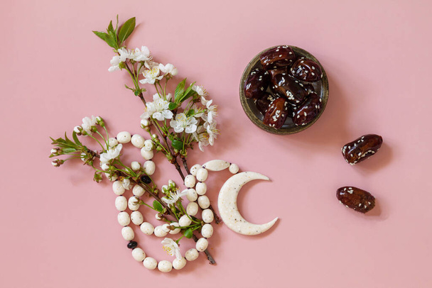 Ramadan food and drinks concept. Ramadan Islamic rosary beads, white flowers and dates fruit on a light pink background. Iftar dinner. Top view flat lay.  - Photo, Image
