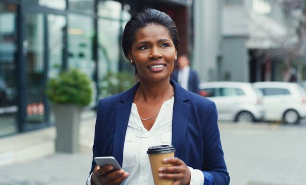 Im well connected in the business world. Shot of an attractive young businesswoman holding her cup of coffee and a cellphone while out in the city. - 写真・画像