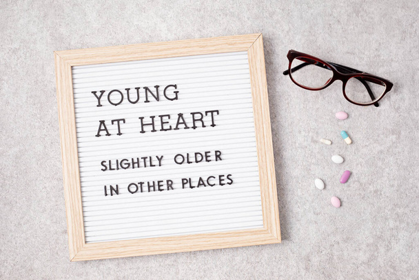 Yüreği genç olan bir mektup tahtası, başka yerlerde biraz daha yaşlı. Arka planda gözlük ve ilaçlar var. Yaşlanma kavramı - Fotoğraf, Görsel