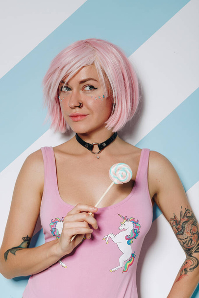Sexy jeune femme tatouée avec la coiffure rose et beau maquillage tenant bonbons sucette colorée douce - Photo, image