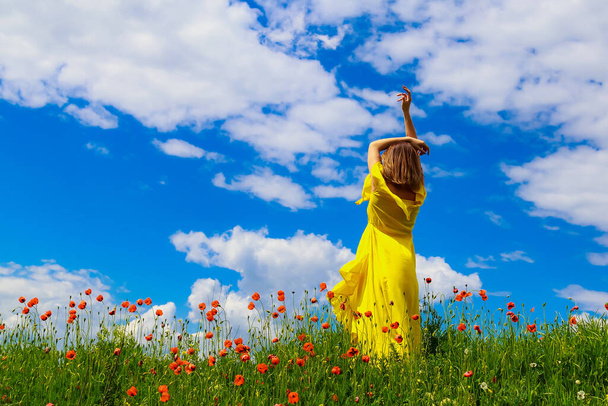 Uma mulher em um vestido amarelo com as mãos levantadas em um campo de papoula contra o fundo de um belo céu. Conceito de liberdade - Foto, Imagem