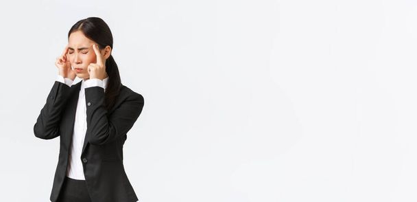 Profil azjatyckiej bizneswoman w czarnym garniturze o migreny, dotykając świątyń i grymaszenie od bolesnego uczucia w głowie. Kierowniczka biura ma bóle głowy, potrzebuje środków przeciwbólowych, białe tło - Zdjęcie, obraz